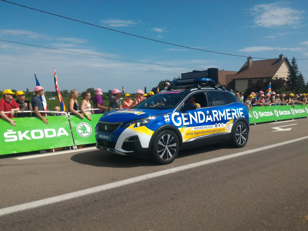 Polizeifahrzeug fährt an einer Menge mit Fahnen und einem Banner vorbei, auf dem "Gendarmerie de la Tour de France" steht, mit Bäumen, Gebäuden und einem bewölktem Himmel im Hintergrund.