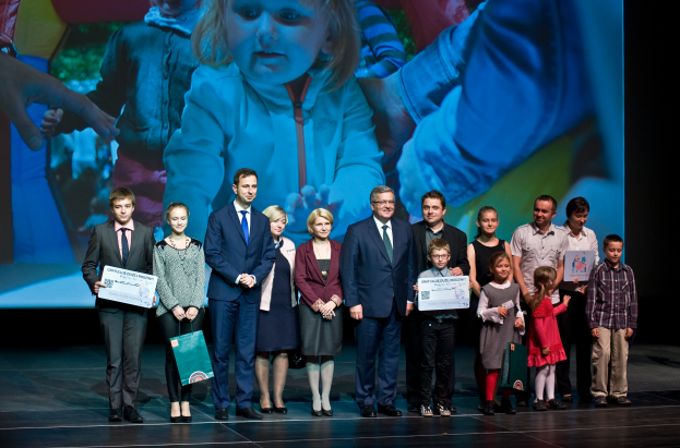 Eine Gruppe von Menschen auf einer Bühne, einige halten Taschen und Urkunden, im Hintergrund ein Bildschirm mit einem Kind und anderen Menschen.