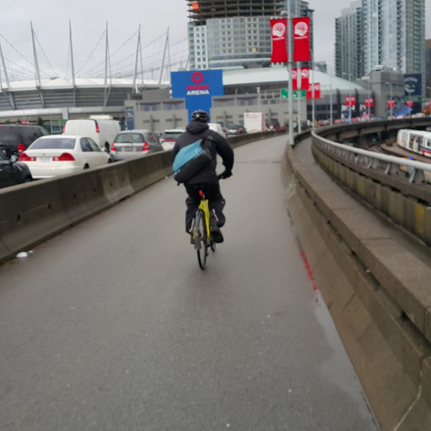 Eine Person mit Helm und Rucksack sitzt auf einem Fahrrad auf einer Straße mit parkenden Autos und Gebäuden im Hintergrund, mit Schildern an Pfosten in der Nähe.