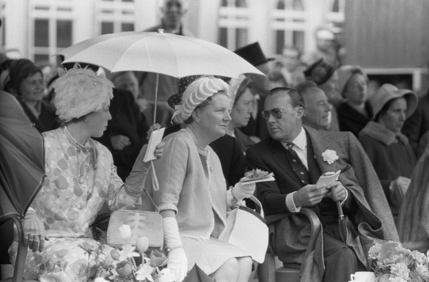 Schwarzes und weißes Foto von Queen Elizabeth II. und Prinz Philip, die auf Stühlen mit Schirmen sitzen, umgeben von Menschen mit Hüten und Büchern, mit einem Gebäude und Blumenschmuck im Hintergrund.