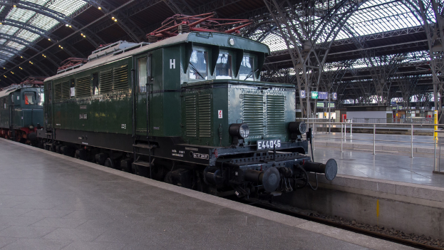 Ein Zug auf Schienen mit Geländern und Tafeln im Hintergrund, Lampen auf dem Dach und Himmel darüber, mit einem Boden darunter.