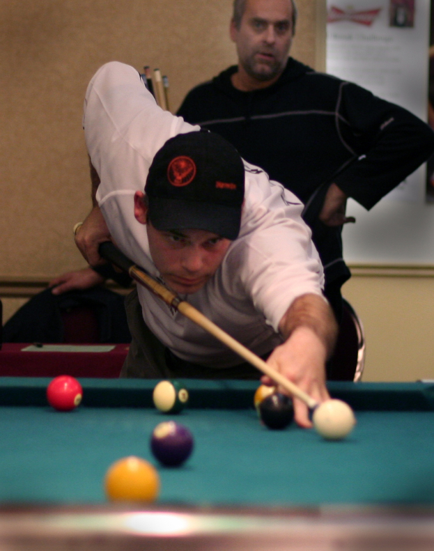 Ein Mann in einem weißen T-Shirt und schwarzer Mütze spielt Snooker, hinter ihm steht ein Mann in einem schwarzen T-Shirt und im Hintergrund ist eine weiße Tafel zu sehen.