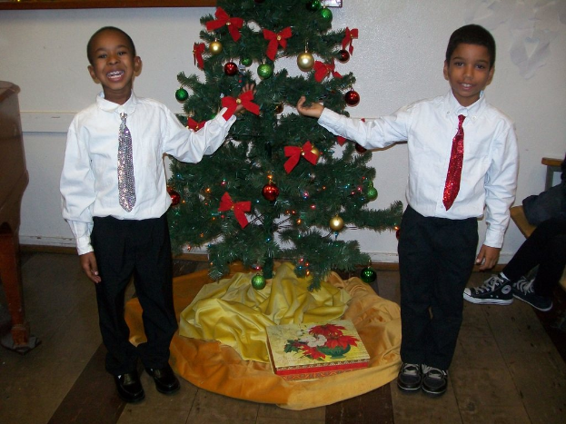 Ein geschmückter Weihnachtsbaum steht in einem Raum mit einer Kiste und anderen Gegenständen auf dem Boden, einem hölzernen Tisch links, einem Stuhl rechts und zwei Jungen in weißen Hemden, die strahlen und den Baum halten.