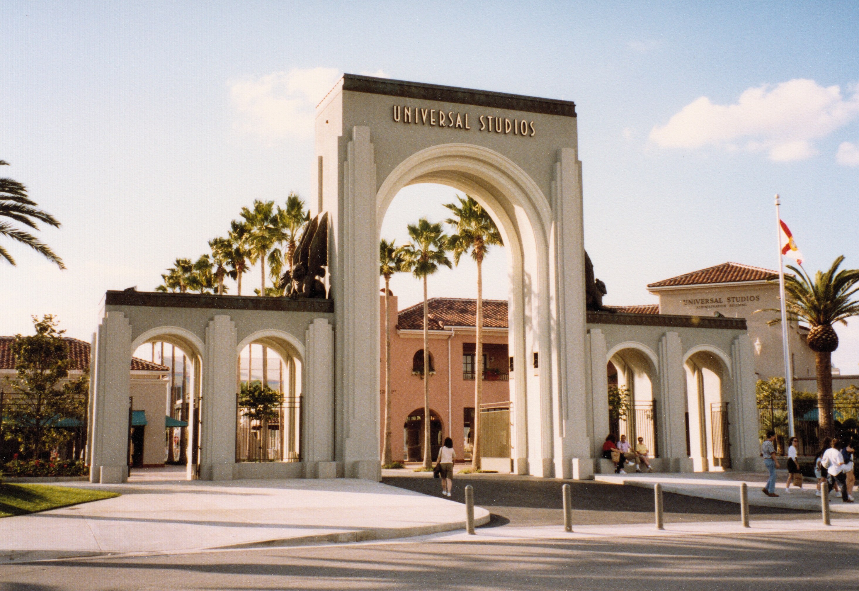 Eingang zum Universal Studios Hollywood mit einem Tor mit Text, einer Statue, Menschen, Bäumen, Gebäuden, einem Fahnenmast mit einer Fahne und einem bewölkten Himmel.
