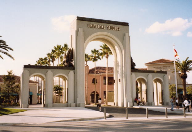 Eingang zum Universal Studios Hollywood mit einem Tor mit Text, einer Statue, Menschen, Bäumen, Gebäuden, einem Fahnenmast mit einer Fahne und einem bewölkten Himmel.