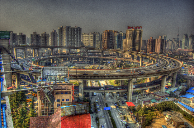 Eine Stadtansicht mit einer zentralen Ringstraße mit Fahrzeugen, einer unteren Straße mit Autos und Fahrrädern, zahlreichen hohen Gebäuden im Hintergrund, einem Himmel oben, Bäumen auf der linken unteren Seite und kleinen Häusern, die über die Stadt verteilt sind.