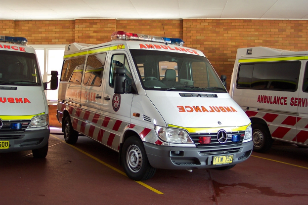 Ein weißer Krankenwagen steht in der Mitte des Bildes, im Hintergrund eine Backsteinwand.