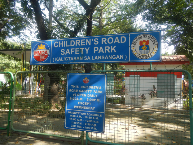 Metallzaun um einen Kinderverkehrssicherheitspark mit zwei blauen Informationsschildern, einem kleinen Haus mit Dach, Pflanzen, einer Baumgruppe und einem bewölkten Himmel.