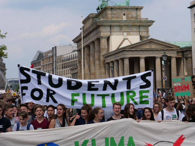 Eine Gruppe von Schülern marschiert in Berlin, die eine bunt bemalte "Students for Future"-Schriftzug-Tafel trägt, vor einer Kulisse aus Gebäuden, Bäumen und Himmel.
