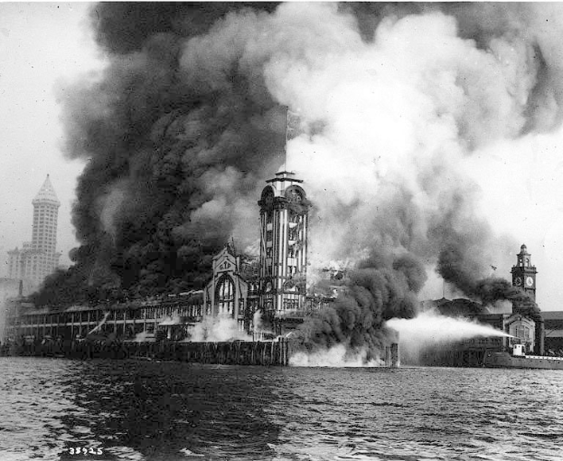 Schwarzes und weißes Foto eines großen brennenden Gebäudes mit Rauch, der herausquillt, einem Gewässer im Vordergrund und anderen Gebäuden und Himmel im Hintergrund.