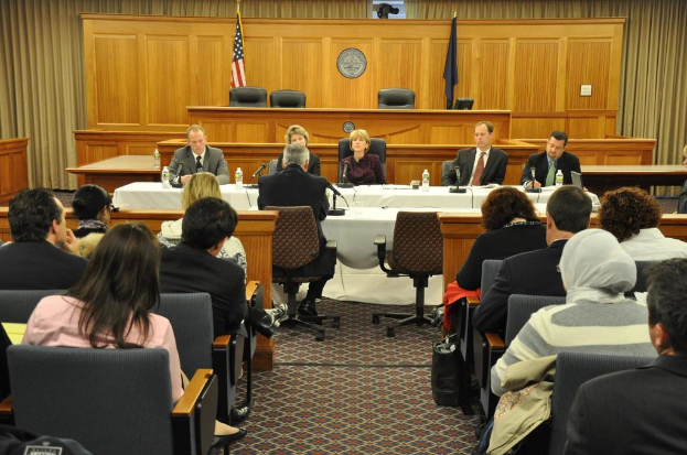 Ein Versammlungsraum mit einer Gruppe von Menschen, die auf Stühlen sitzen, zwei ausländischen Flaggen, einem orangen Schreibtisch, Wasserflaschen und einem Mikrofon auf dem Schreibtisch.