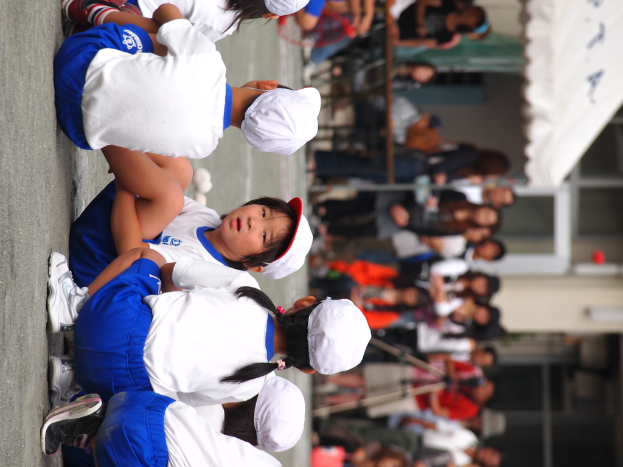 Kinder auf der Straße sitzend, im Hintergrund Menschen stehend, ein Stativ, ein Zelt und ein Gebäude mit Glaswänden.