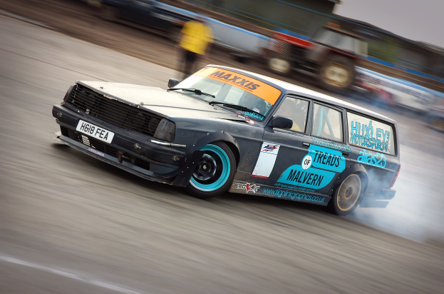 Ein Auto, das auf einer Rennstrecke fährt und Rauch ausstößt, umgeben von anderen Fahrzeugen und Zuschauern, mit einem unscharfen Hintergrund, der hohe Geschwindigkeit andeutet.