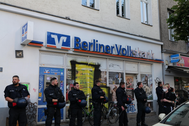 Gruppe von Polizeibeamten vor einer Berliner Volksbank stehend, einige mit Helmen, mit einem Auto auf der rechten Seite und einem Baum auf der linken Seite, Gebäude mit Fenstern und Texttafeln im Hintergrund.