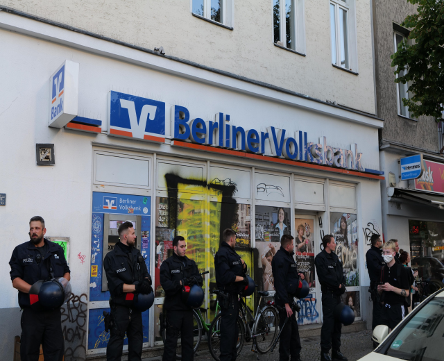 Gruppe von Polizeibeamten vor einer Berliner Volksbank stehend, einige mit Helmen, mit einem Auto auf der rechten Seite und einem Baum auf der linken Seite, Gebäude mit Fenstern und Texttafeln im Hintergrund.