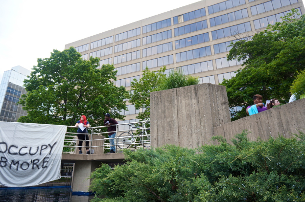 Menschen auf einer Brücke mit einem Banner und einem Fahrrad, darunter Wasser, mit Bäumen, Gebäuden und Himmel im Hintergrund.