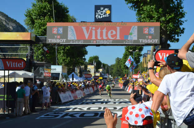 Eine Gruppe von Radfahrern fährt auf einer Straße während der Tour de France 2016, mit Zuschauern auf beiden Seiten, Bäumen, einem Hügel und einem klaren blauen Himmel im Hintergrund und einem Bogen mit "Tour de France 2016" oben.