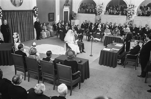 Schwarzes und weißes Foto einer Hochzeitszeremonie in einem großen Raum mit sitzenden und stehenden Gästen, Tischen mit Mikrofonen und Papieren, einer Wand mit einem Fotorahmen und Blumenbouquets, einem Vorhang und einem Podium mit einem Mikrofonständer.