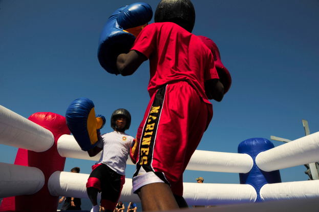 Zwei Boxer in einem Boxring, einer in rotem Hemd und blauen Shorts, kämpfen mit Handschuhen, umgeben von ein paar Leuten und unter einem klaren blauen Himmel.