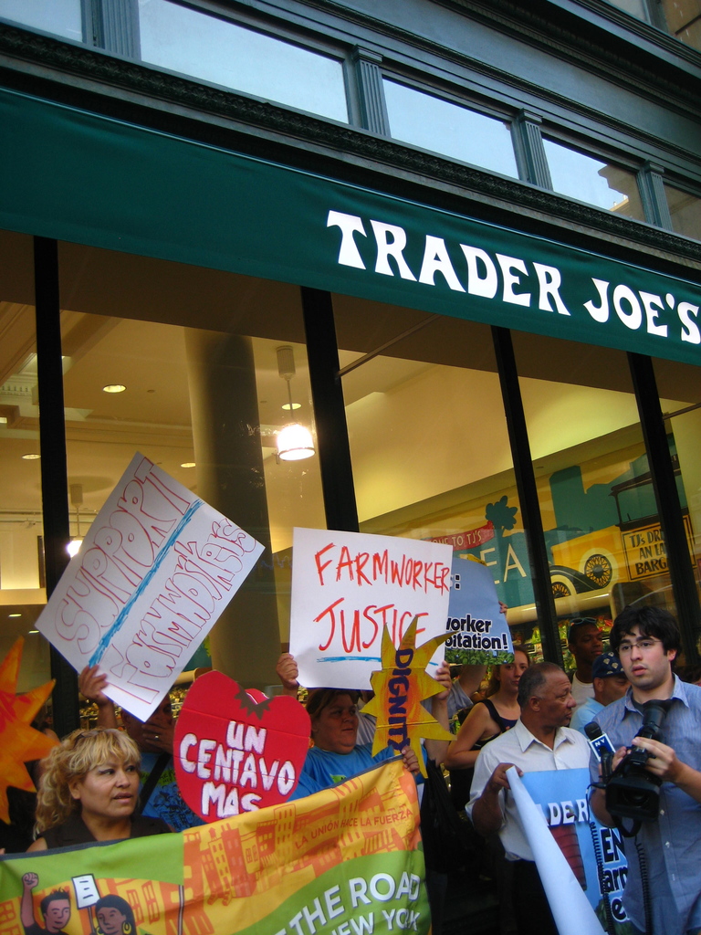 Eine Gruppe von Menschen steht in der Nähe eines Ladens und hält Schilder und Plakate in den Händen, wobei hinter ihnen eine Säule und Lichter auf dem Dach zu sehen sind.