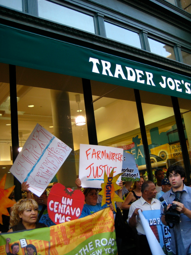 Eine Gruppe von Menschen steht in der Nähe eines Ladens und hält Schilder und Plakate in den Händen, wobei hinter ihnen eine Säule und Lichter auf dem Dach zu sehen sind.