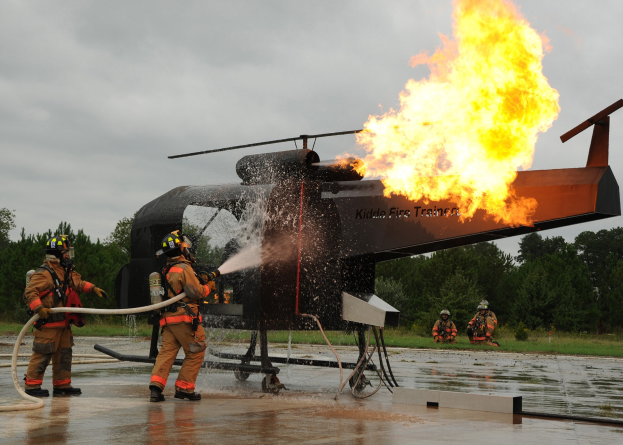 Feuerwehrleute in Helmen spritzen Wasser auf einen brennenden Hubschrauber, während im Hintergrund Bäume und ein klarer blauer Himmel zu sehen sind.