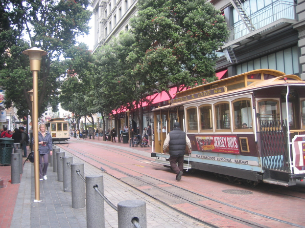 Ein Zug auf Schienen mit Text an der Seite, eine Person in der Nähe, kleine Pfosten mit Seilen auf der linken Seite, Straßenlaternen, Bäume, viele Menschen, ein Müllcontainer auf der linken Seite und Gebäude mit Leitern auf der rechten Seite.