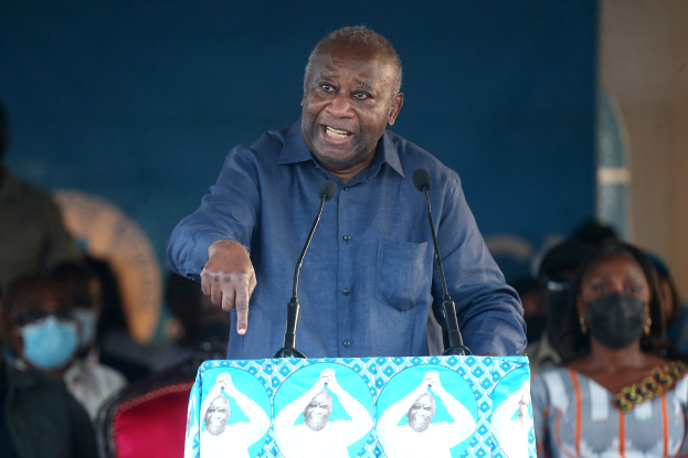 Ein Mann in einem blauen Hemd spricht in zwei Mikrofone an einem Rednerpult, mit einer Menge und sitzenden Personen, einige tragen Masken, im Hintergrund.