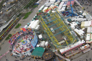 Luftaufnahme eines Vergnügungsparks mit einem zentralen Riesenrad, umgeben von verschiedenen Attraktionen, Gebäuden, Fahrzeugen, Menschen, Straßen und Grünflächen.