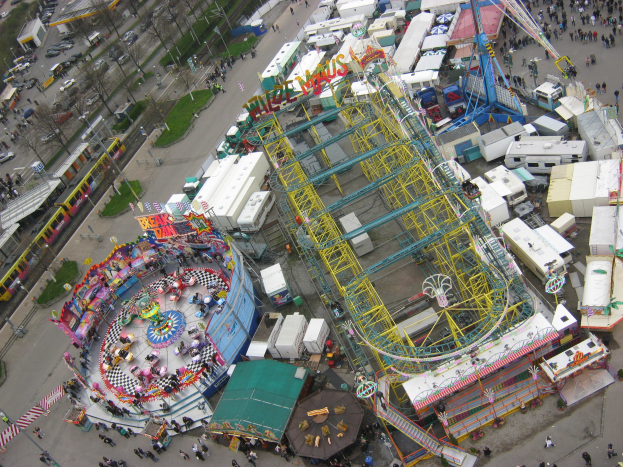 Luftaufnahme eines Vergnügungsparks mit einem zentralen Riesenrad, umgeben von verschiedenen Attraktionen, Gebäuden, Fahrzeugen, Menschen, Straßen und Grünflächen.